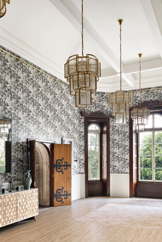 A spacious room for international students, featuring tall windows, patterned grey wallpaper, three large gold pendant lights, wooden flooring, a geometric sideboard, ceramic vase, and decorative wooden screen. Trees are visible outside.